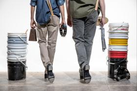 2 tap dancers, each with a pair of drumsticks in their back pocket. One is holding a cowbell and tambourine, the other holding a cymbal on a stand. 2 stacks of buckets ready to be drummed upon.