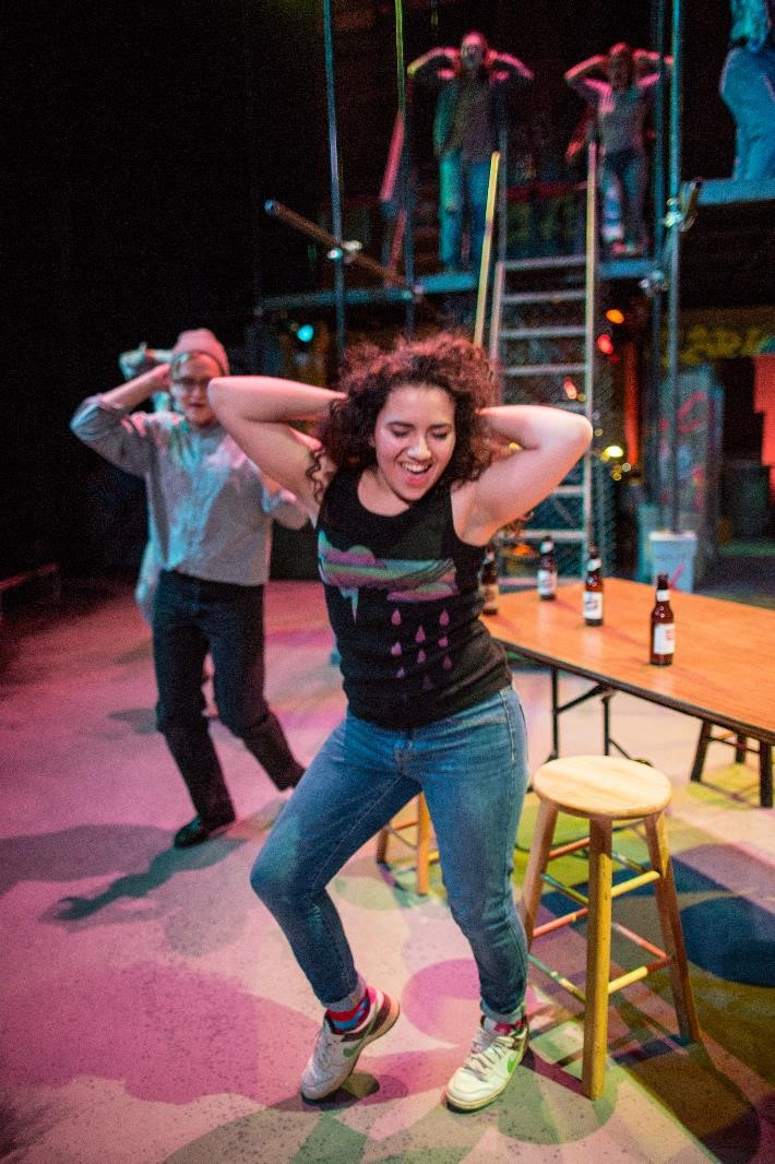 Laila stands in the foreground, dancing. Her hands are behind her head and her right knee is bent. She wears cuffed jeans, multi-colored socks, white and brown sneakers, and a black tank top with a multi-colored storm cloud on it.