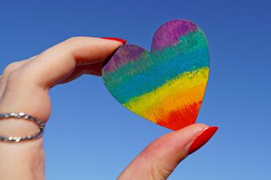 A hand with red painted fingernails holds up a pride heart