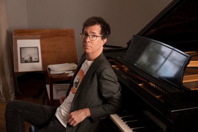 Photograph of Ben Folds seated on a piano bench leaning backwards onto an open grand piano keyboard. 