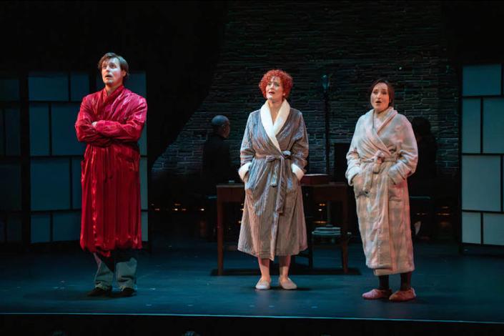 Three actors stand on stage in a line against a dark background. On the left is a man in a long red robe. In the middle is a woman in a long grey robe. On the right is a woman in a light grey robe.