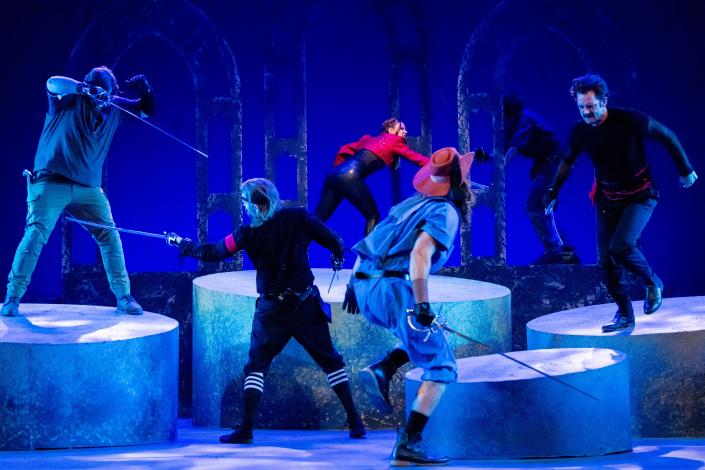 a collection of swordfighting actors on a blue-lit stage littered with a series of cylindrical platforms.