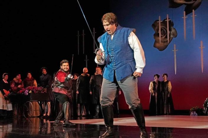 An opera performer, dressed as a young man, who is passionately singing about loss while beginning to raise a sword up in vengeance. In the background is an opera chorus dressed as townsfolk, along with a performer dressed as a royal wielding a sword and staring at the first young man with violent glee. The photo was taken by Ken Howard.