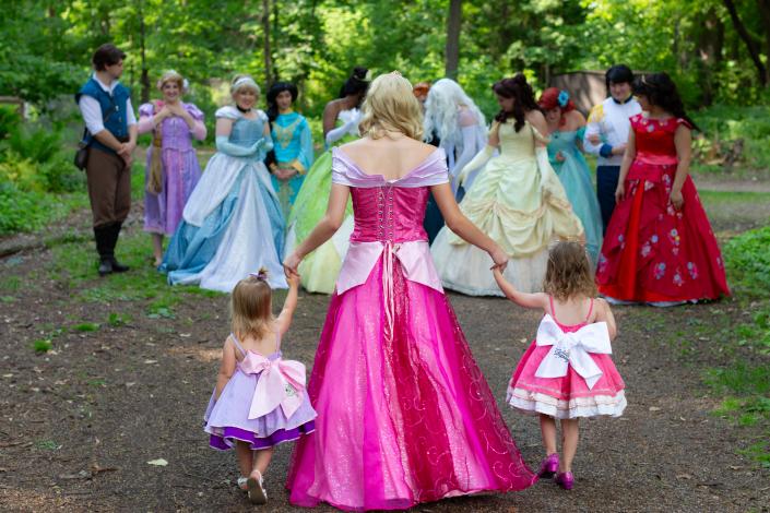 Princess in pink walks holding the hands of two little girls on each side, one in pink and one in purple, toward a large group of princesses.