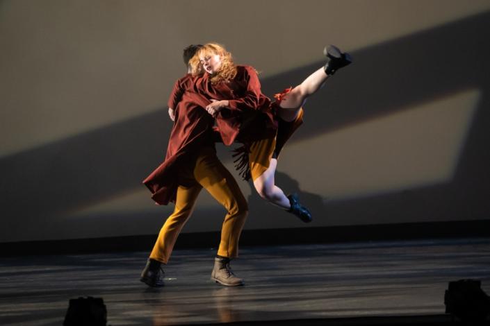 Two dancers in gold pants and red jackets. One lifting the other on a gray stage background. 