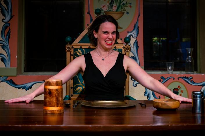 A woman with a wicket grin spreads her arms out in invitation, behind a rustic table set for dinner.