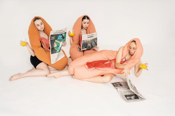 A photo of three femme people in vulva costumes lounging and reading newspapers