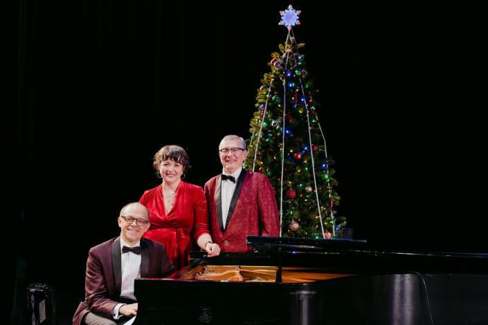 Portrait of three actors surround a grand piano with a decorated tree in the background. 