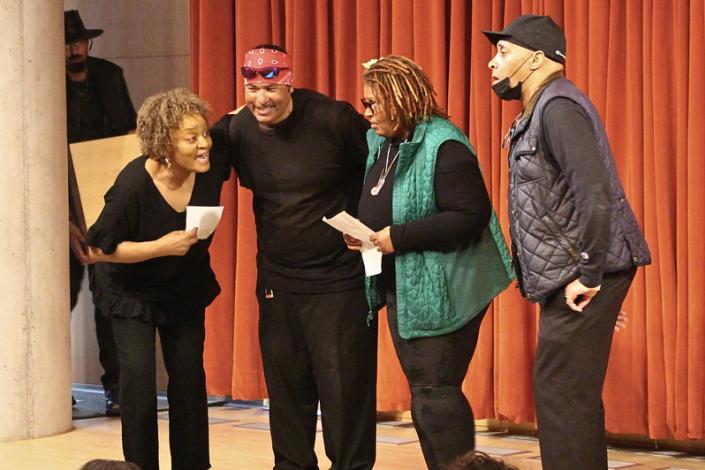 Four performers singing together, wearing black and standing in front of a red curtain.