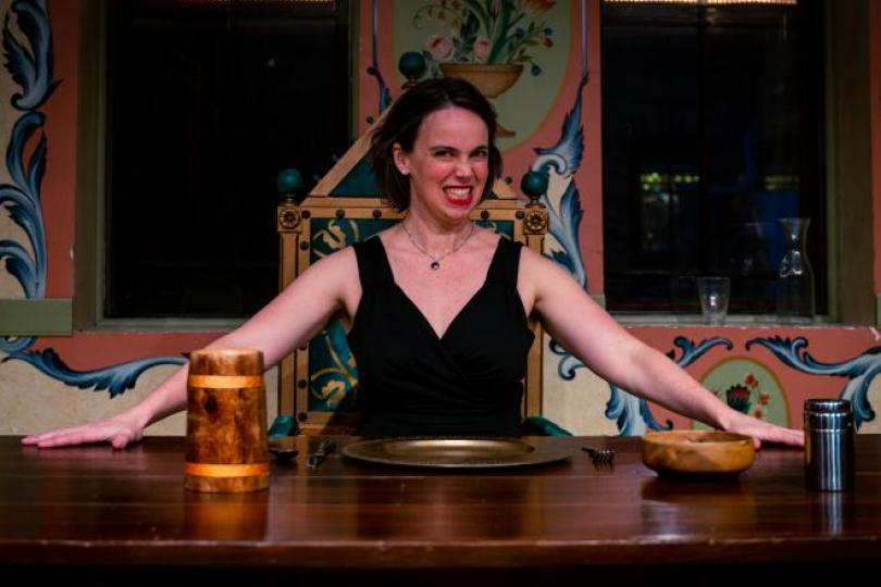 Woman sitting behind large wooden table in black dress giving audience a sinister smile.