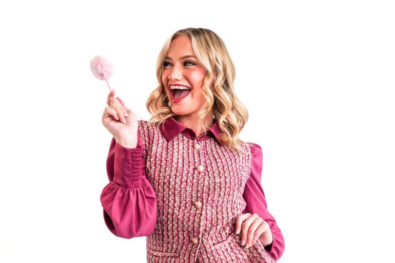 A blonde woman stands smiling in a pink shirt and vest holds up a pink pompom pen.